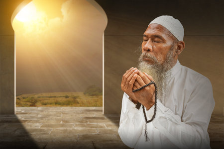Muslim man with a beard wearing a white cap praying with prayer beads on his hands inside the mosqueの写真素材
