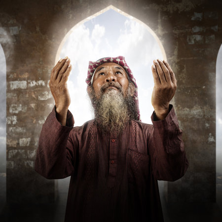 Muslim man with a beard wearing keffiyeh with agal in praying while raised hands on the mosqueの写真素材