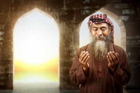 Muslim man with a beard wearing keffiyeh with agal in praying while raised hands on the mosqueの写真素材