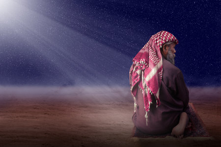 Muslim man with a beard wearing keffiyeh with agal in praying position (salat) on the prayer rug with the night scene backgroundの写真素材