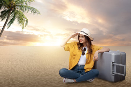 Asian woman with a hat and suitcase sitting while looking at something on the beach with a sunset scene background. World tourism day conceptの写真素材