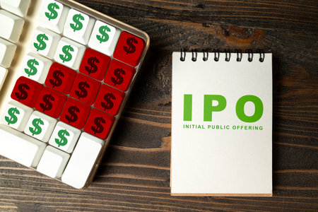 Closeup view of a notebook with IPO text and a computer keyboard with a currency button on a wooden...