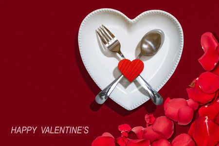 Heart plate with silverware of spoon and fork with a red heart and rose petal over a red background. Romantic dinner Valentine's Day conceptの写真素材