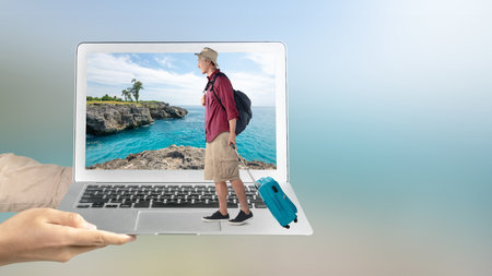 A human hand holding a laptop with an Asian man with a hat and a backpack carrying a suitcase standing with a sea cliff and an ocean view on the laptop screen. Traveling conceptの写真素材