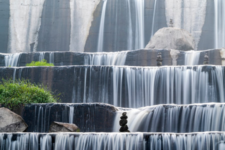 A waterfall with rocks and a small plant in the foreground. The water is flowing down the rocks and the plant is growing in the water in Grojogan Watu Purbo, Yogyakarta, Indonesiaの写真素材