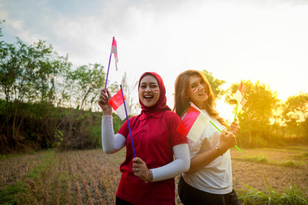 Portrait of two Indonesian women with excited expressions holding small Indonesian flag outdoors with sunrise sky backgroundの写真素材