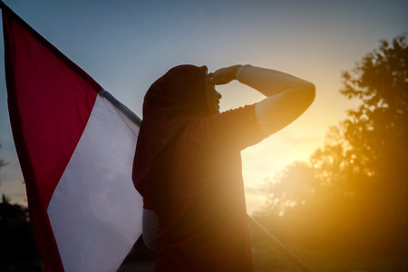 Portrait of a woman holding an Indonesian flag with respectful gesture outdoors with a sunrise sky backgroundの写真素材
