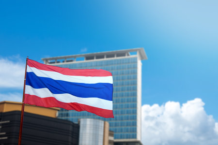 A Thailand flag with the colors red, white, and blue is flying in front of a large buildingの写真素材