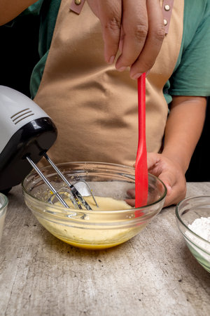 A person is mixing ingredients in a bowl with a red spatula. The person is wearing an apron and is...