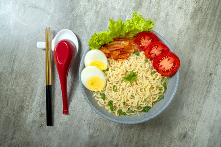 A bowl of instant noodles with eggs and tomatoes. The bowl is on a table with chopsticks and a spoon