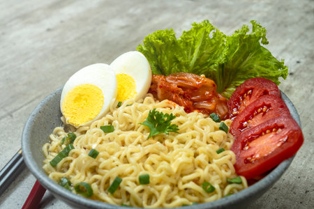A bowl of instant noodles with eggs and tomatoes. The bowl is on a table with chopsticks and a spoon