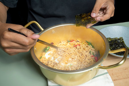 A person is eating instant noodles with chopsticks. The noodles are in a bowl and the person is...