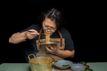 A man is eating instant noodles from a bowl. She is wearing an apron and has a spoon in her mouthの写真素材