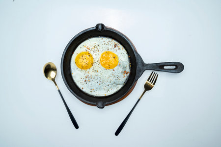 A pan of sunny side up eggs and a fork and spoon on a white backgroundの写真素材