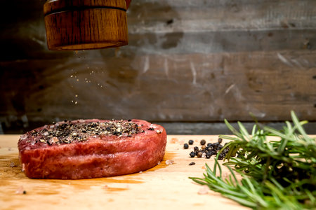 A pepper grinder is on a wooden cutting board with a piece of tenderloin meat and some herbs