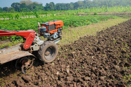 A tractor is working in a field with dirt and grass. The tractor is red and orange. The sky is blue...
