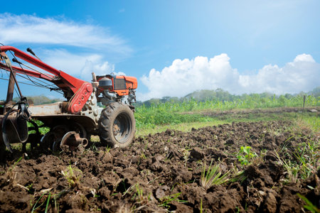 A tractor is working in a field with dirt and grass. The tractor is red and orange. The sky is blue and there are clouds in the backgroundの写真素材