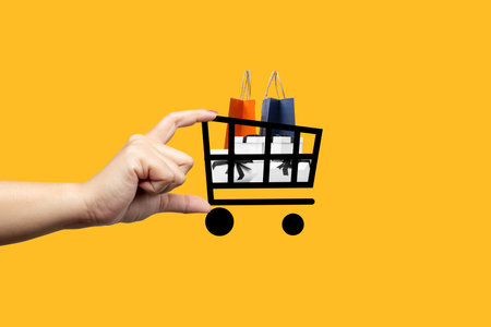 A hand holding a shopping cart with two bags on it. The cart is drawn in black and white and the bags are orange and blue. Collage artの写真素材