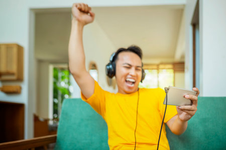 An Indonesian man in a yellow shirt is playing a video game on a phone while wearing headphones. He...