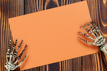 A human skeleton hand holding an orange piece of paper on a wooden background. The blank orange piece of paper for copy space. The image has a creepy and unsettling mood, a horror or Halloween decorationの写真素材