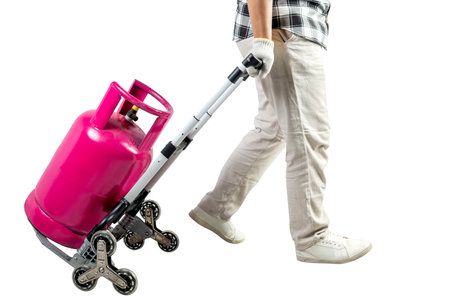 A man is carrying a pink gas cylinder on a cart isolated over a white background. The man is wearing a plaid shirt, gloves, and white pantsの写真素材