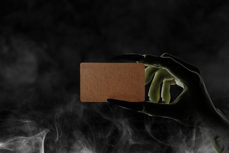 Witch hand with long black nail holding piece of brown paper with smoke on dark background. Blank piece of brown paper for copy space. Evil, satan, zombie or monster hand. Spooky Halloween conceptの写真素材