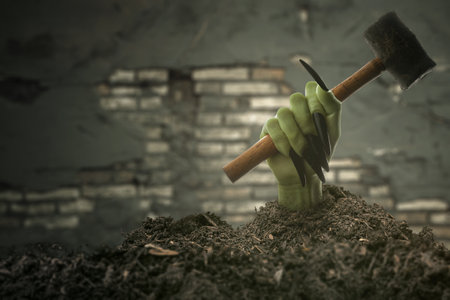 Witch hand with long black nail coming out from grave ground holding a hammer with brick wall...