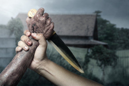 Zombie hand with bloody wound fighting with man on midnight background. Zombie hand holding knife is pointed at the man hand, creating tense and dangerous atmosphere. Spooky Halloween conceptの写真素材