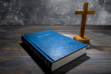 Wooden religion cross and blue book with the word Holy Bible. It is laying on a wooden table....