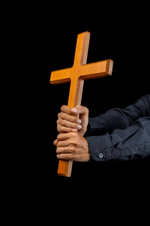 Man holding a wooden religion cross in his hand on black background. Symbol of faith and worship in...