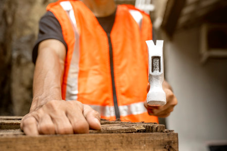 Workers in safety vest using hammer to to work assembling pallet parts. The scene is set in a warehouse background. Woodworking factoryの写真素材