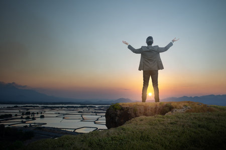 Rear view of businessman standing while looking at mountain view with raised arm on top of valley edge. Business, success, and motivation conceptの写真素材