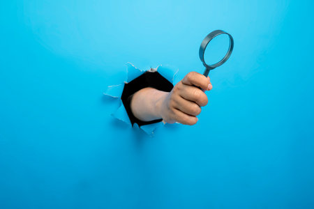 Human hand holding magnifying glass through ripped paper. Mockup or Copy space. Concept of Business or New Ideasの写真素材