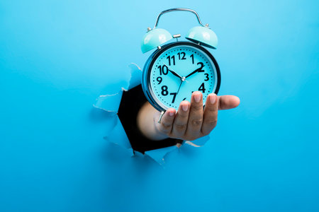 Human hand holding alarm clock through ripped paper. Mockup or Copy space. Concept of time...