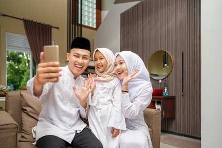 Portrait of the Indonesian Muslim family with mobile phone taking selfie or video call while celebrating Eid Mubarak. Concept of Happy Lebaran, Eid Al Fitr, Idul Fitri, and Ramadan Mubarak celebrationの写真素材