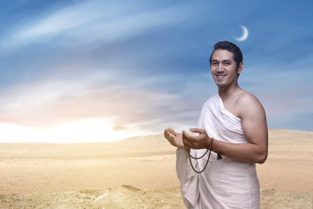 Indonesian southeast asian muslim man wearing white traditional clothes or ihram raises his hand and prays while holding prayer beads on the hajj. Concept of hajj or umrahの写真素材