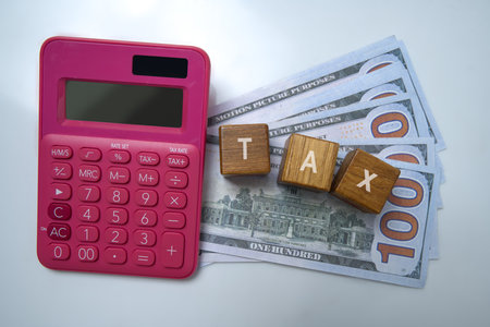 Top view of pink calculator and wooden cubes with tax text and a pile of 100 dollar banknotes on a white background. Concept of finance and taxationの写真素材