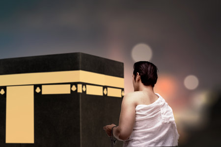Rear view of an Indonesian southeast asian muslim man wearing white traditional clothes or ihram raises his hand and prays while holding prayer beads in front of the Kaaba. Concept of hajj or umrahの写真素材