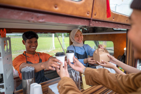 Indonesian southeast asian male and a Chinese muslim female in hijab couple baristas serving drinks and snacks to customers. A small business of a coffee shop or cafe on a truckの写真素材