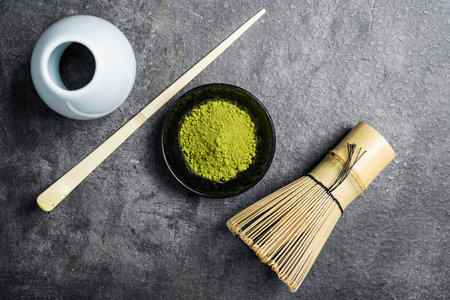 Matcha green tea powder in the black bowl and a traditional bamboo brush whisk with a spoon on a black table. Accessories for making matcha green tea. Top viewの写真素材
