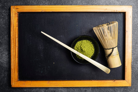 Matcha green tea powder in the black bowl and a traditional bamboo brush whisk with a spoon on a black table. Accessories for making matcha green tea. Copy space. Top viewの写真素材