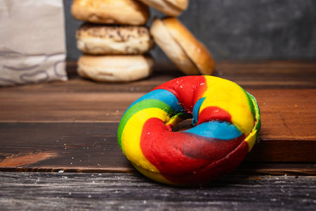 Closeup view of a rainbow bagel bread and a heap of a collection of bagel breads in various kinds with a brown paper bag on a wooden boardの写真素材