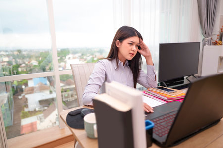 Indonesian southeast asian businesswoman experiencing stress and burnout while working on laptop in hybrid workplace. Paperwork and a cellphone on the table. Business concept. Remote working from homeの写真素材