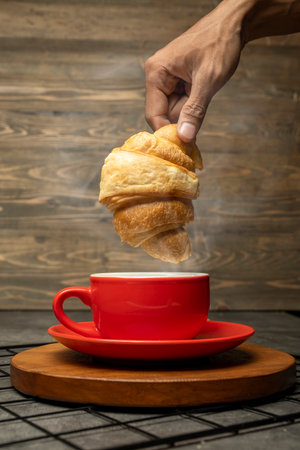 Closeup view human hand dipping a freshly baked croissant into a cup of coffee on a wooden tray with a wooden backgroundの写真素材