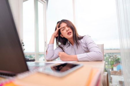 Indonesian southeast asian businesswoman experiencing stress and burnout while working on laptop in hybrid workplace. Paperwork and a cellphone on the table. Business concept. Remote working from homeの写真素材