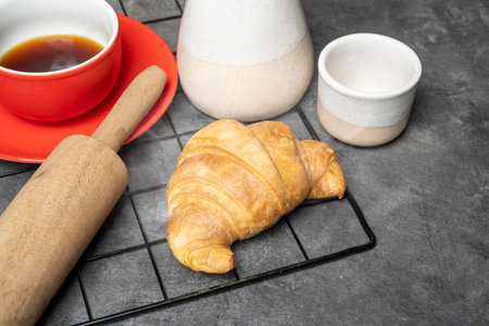 Freshly baked croissant and a cup of coffee with a rolling pin on a cooling tray on the tableの写真素材