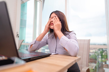 Indonesian southeast asian businesswoman experiencing stress and burnout while working on laptop in hybrid workplace. Paperwork and a cellphone on the table. Business concept. Remote working from homeの写真素材