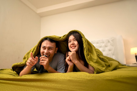 Indonesian southeast asian couple lying down under a blanket while watching a movie on television...