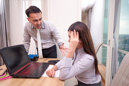 Indonesian southeast asian businessman scolding his frustrated employee for being late, pointing at his watch, the stress of deadlines. Laptop and paperwork on the table. Business conceptの写真素材
