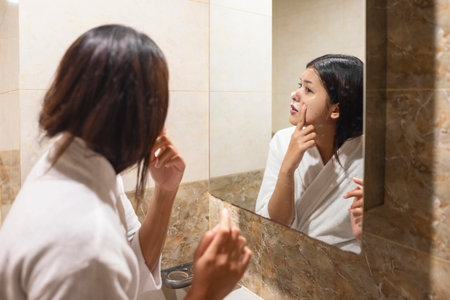 Indonesian southeast asian woman wrapped in a towel, looking at herself in the mirror while squeezing an acne spot. Natural skincare and wellness conceptの写真素材